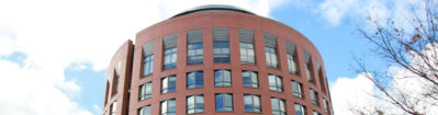 A red brick building with a cylindrical shape, featuring multiple windows in a modern architectural style against a partly cloudy sky.